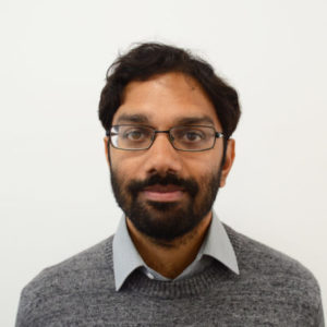 Aveek is a South Asian man with dark brown hair. He is wearing a grey jumper and has on black framed glasses. He is looking at the camera. Aveek is chairing the event. 