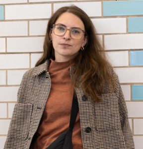 Caroline is a white woman with dark brown hair. She is wearing a rust colour jumper, and wears square-framed glasses. She is looking at the camera.